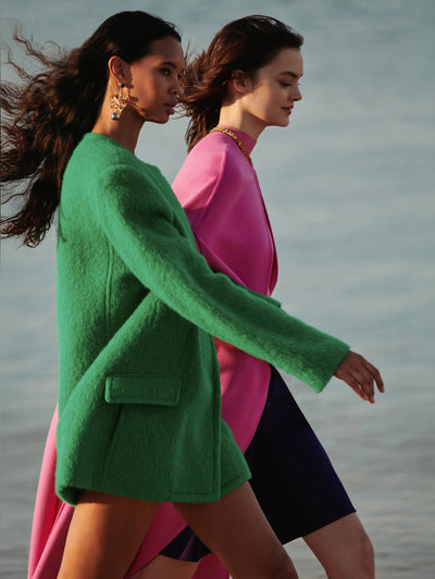Two models walk side-by-side near the ocean in Oscar de la Renta outerwear styles. 
