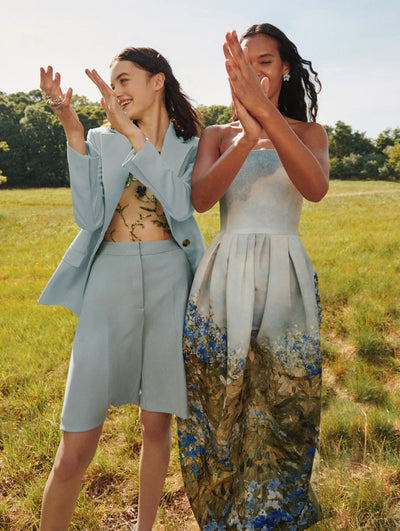 Two models are seen clapping in a field, one wearing an Oscar de la Renta blue blazer and short set and the other wearing an Oscar de la Renta floral strapless gown. 