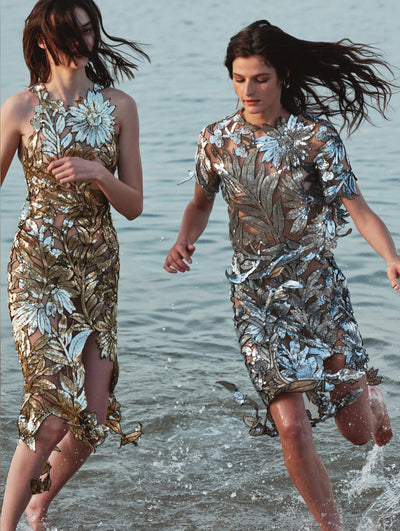 Two models are seen running in the ocean wearing similar laser cut metallic evening dresses. 