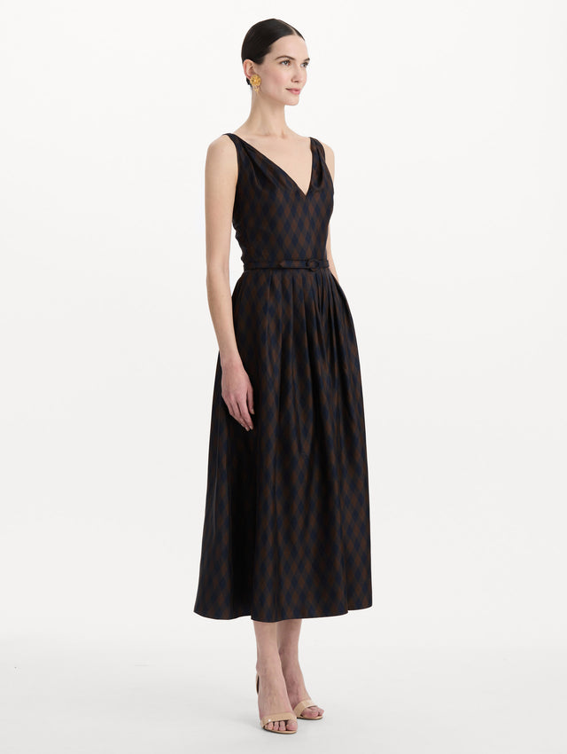 The model is wearing the argyle silk twill dress with a sleeveless silhouette, a V-neckline, and an argyle print in navy and brown. Side view. 