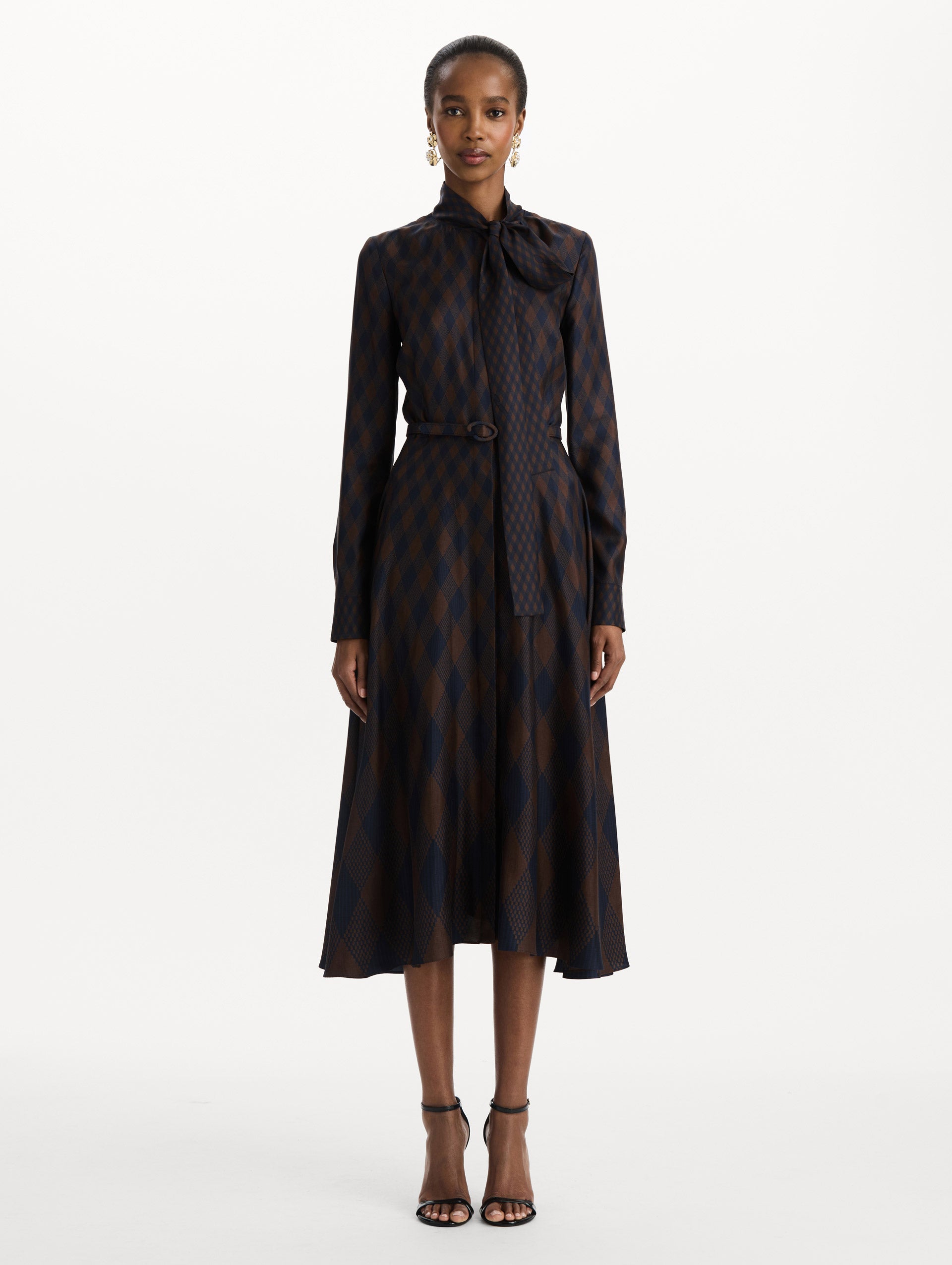 The model is wearing the argyle silk twill tie neck dress with long sleeves, a tie neckline, and an argyle print in navy and brown. Front view. 