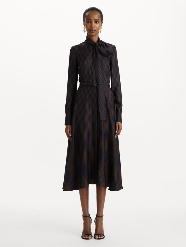 The model is wearing the argyle silk twill tie neck dress with long sleeves, a tie neckline, and an argyle print in navy and brown. Front view. 