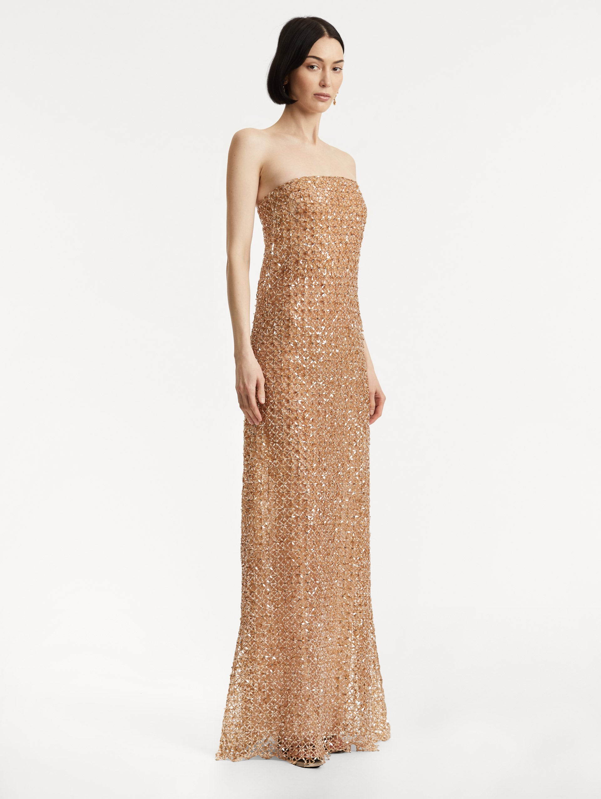 Full body side view of model wearing amber-hued strapless gown with a column silhouette glass floral embroidery in a grid pattern. 