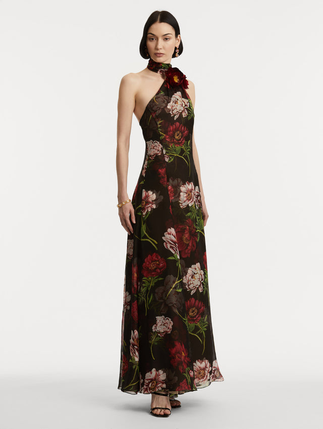 Full body side view of model wearing a silk chiffon gown with black back round and printed pink and red large-scale peonies. A wrap-style halter neckline ties like a scarf, while there is a peony appliqué at the neck