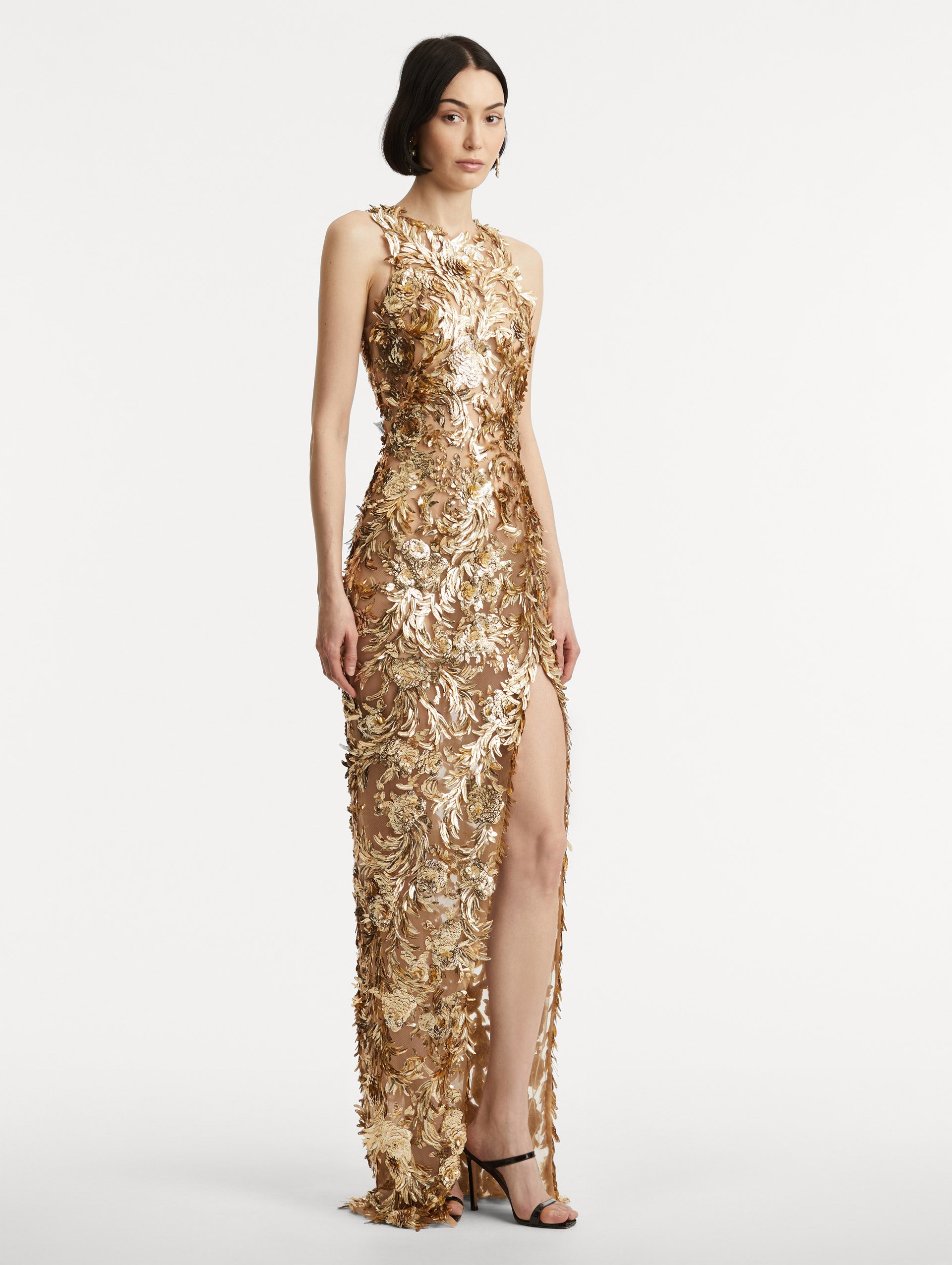 Full body side view of model wearing sleeveless gold-embroidered gown with  a halter neckline and feathers unfurling across a tulle base. Gown features a high center front slit.