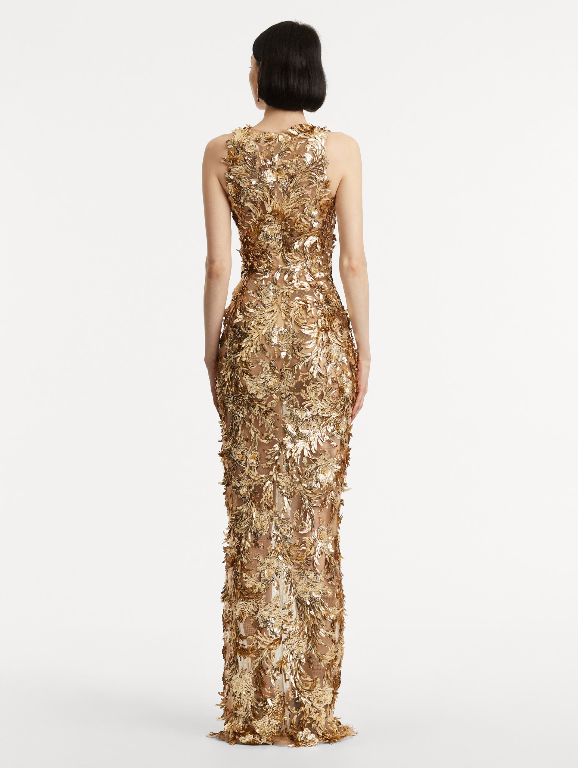 Full body back view of model wearing sleeveless gold-embroidered gown with feathers unfurling across a tulle base. 