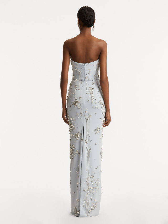 Full body back view of model wearing strapless soft blue gown with a column silhouette and multi-dimensional stones scattered across the dress. Features concealed zipper and central back slit.