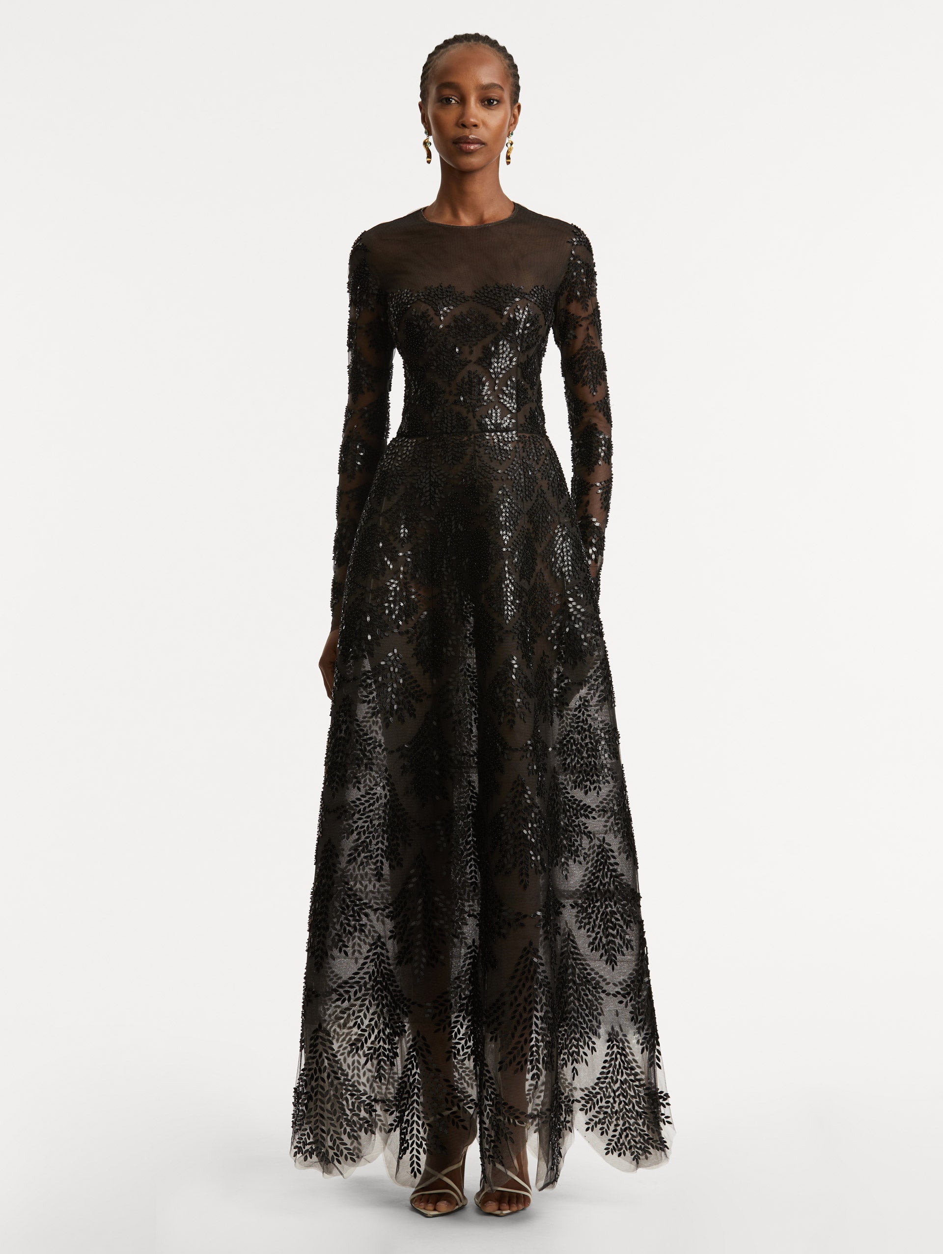 Full body front view of model wearing a black long sleeve gown with an illusion neckline and softly flared skirt. Features black sequin fern embroidery against the tulle base