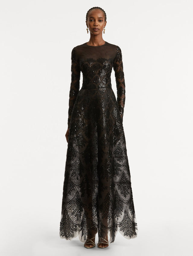 Full body front view of model wearing a black long sleeve gown with an illusion neckline and softly flared skirt. Features black sequin fern embroidery against the tulle base