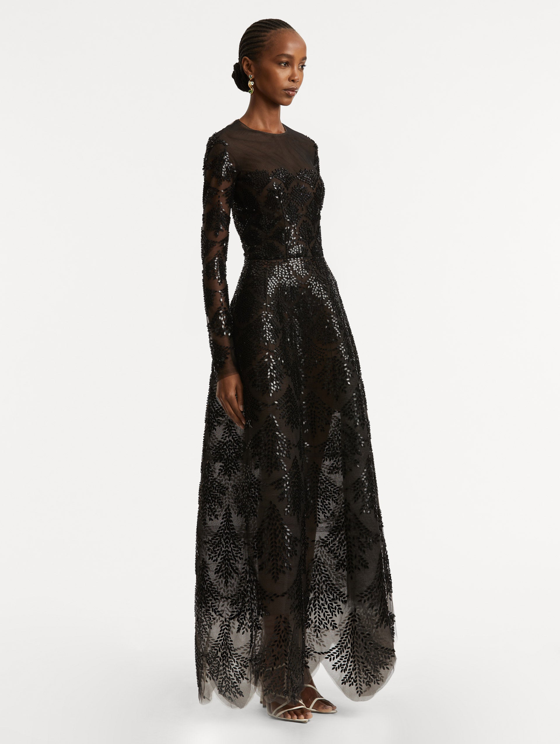 Full body side view of model wearing a black long sleeve gown with an illusion neckline and softly flared skirt. Features black sequin fern embroidery against the tulle base