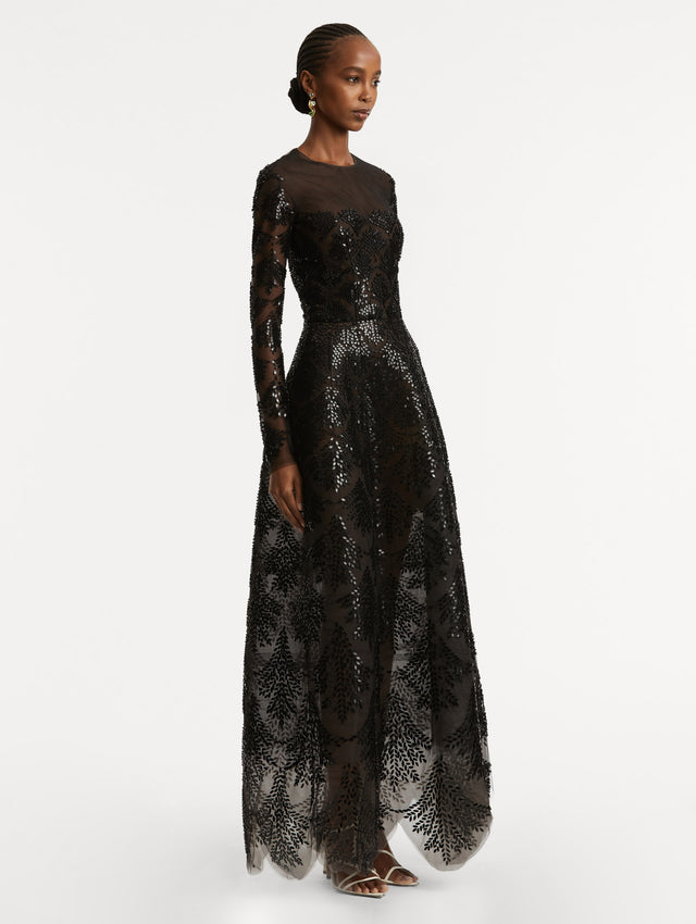 Full body side view of model wearing a black long sleeve gown with an illusion neckline and softly flared skirt. Features black sequin fern embroidery against the tulle base