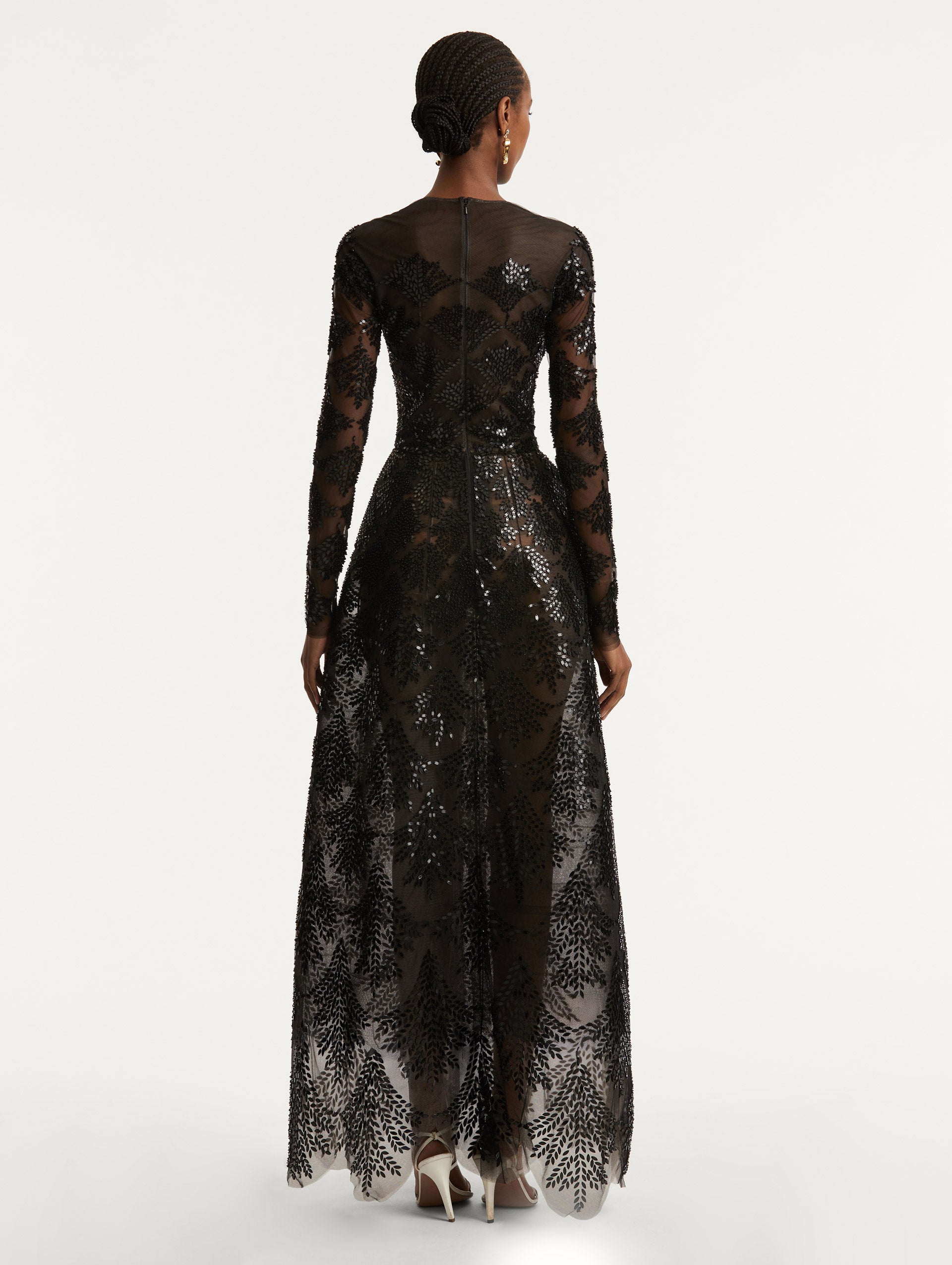 Full body back view of model wearing a black long sleeve gown with an illusion neckline and softly flared skirt. Features black sequin fern embroidery against the tulle base and a concealed black zipper.