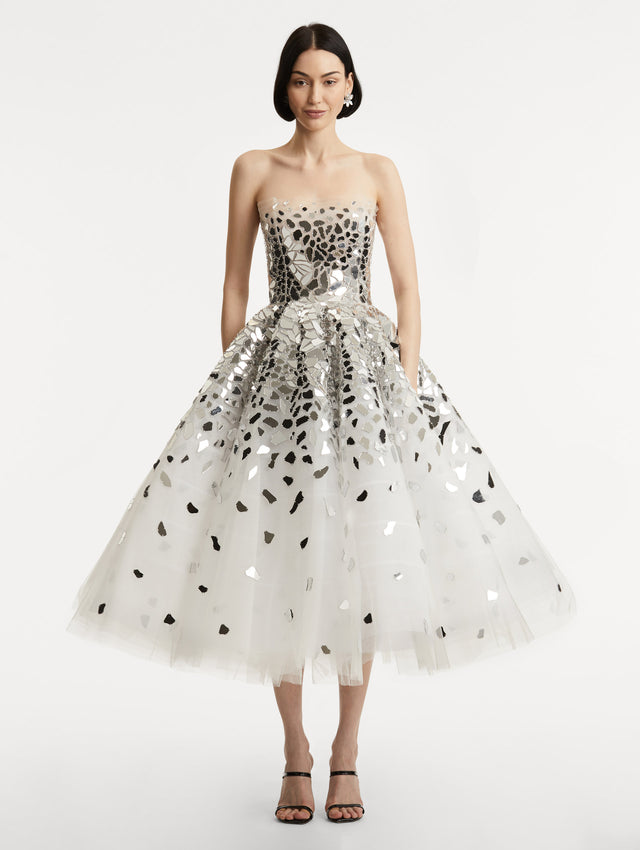 Full body front view of model wearing strapless dress with a white tulle base, mirrored mosaic pieces concentrated on bodice and fade down the skirt