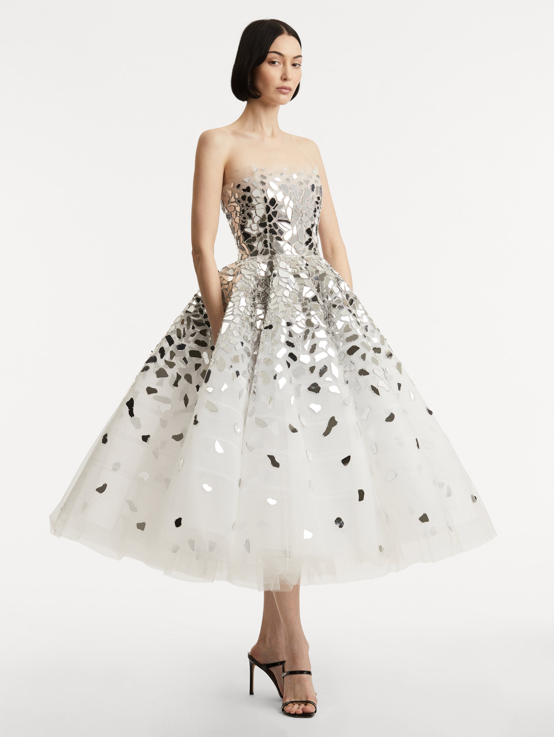 Full body side view of model wearing strapless dress with a white tulle base, mirrored mosaic pieces concentrated on bodice and fade down the skirt