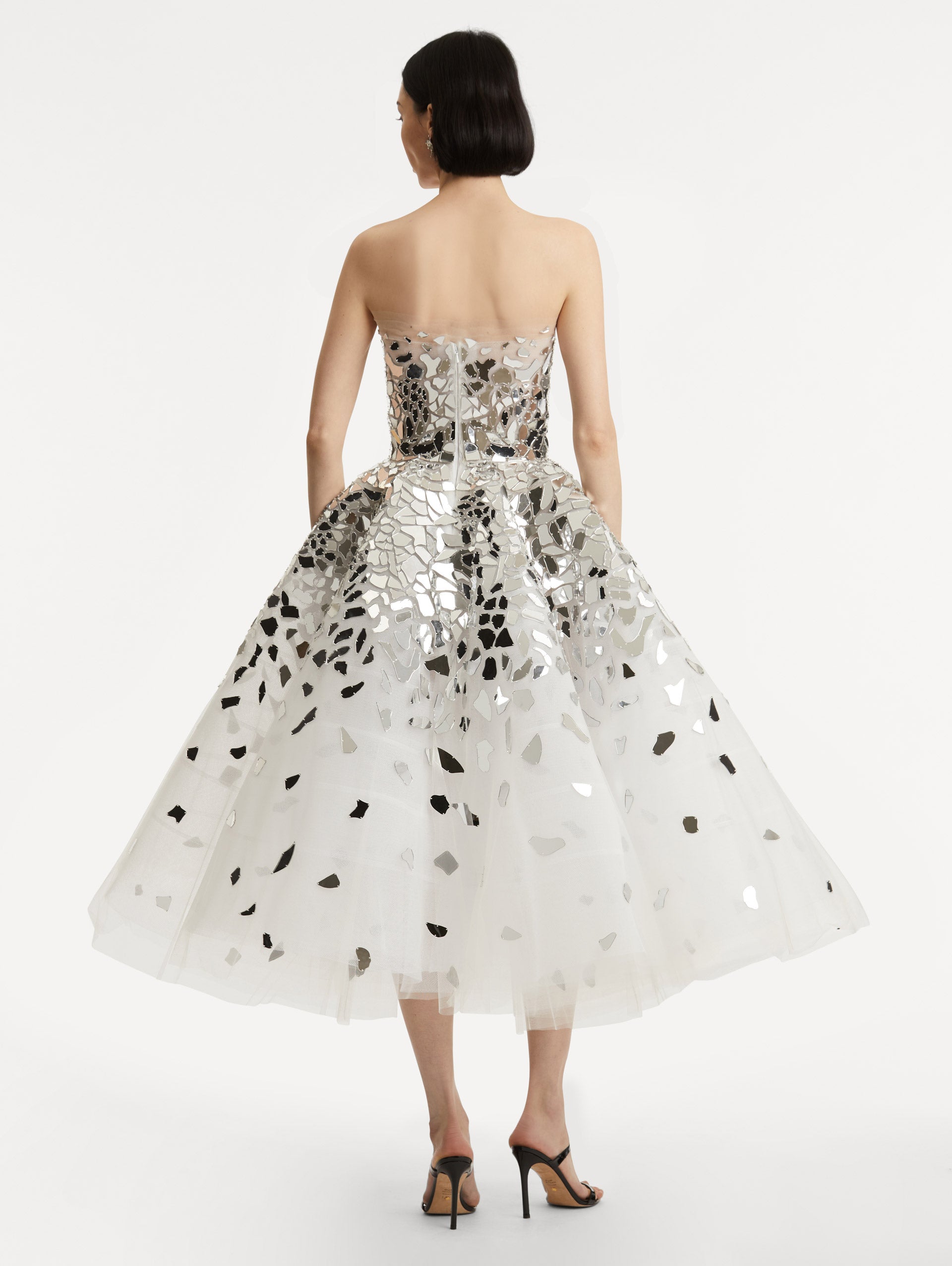 Full body back view of model wearing strapless dress with a white tulle base, mirrored mosaic pieces concentrated on bodice and fade down the skirt