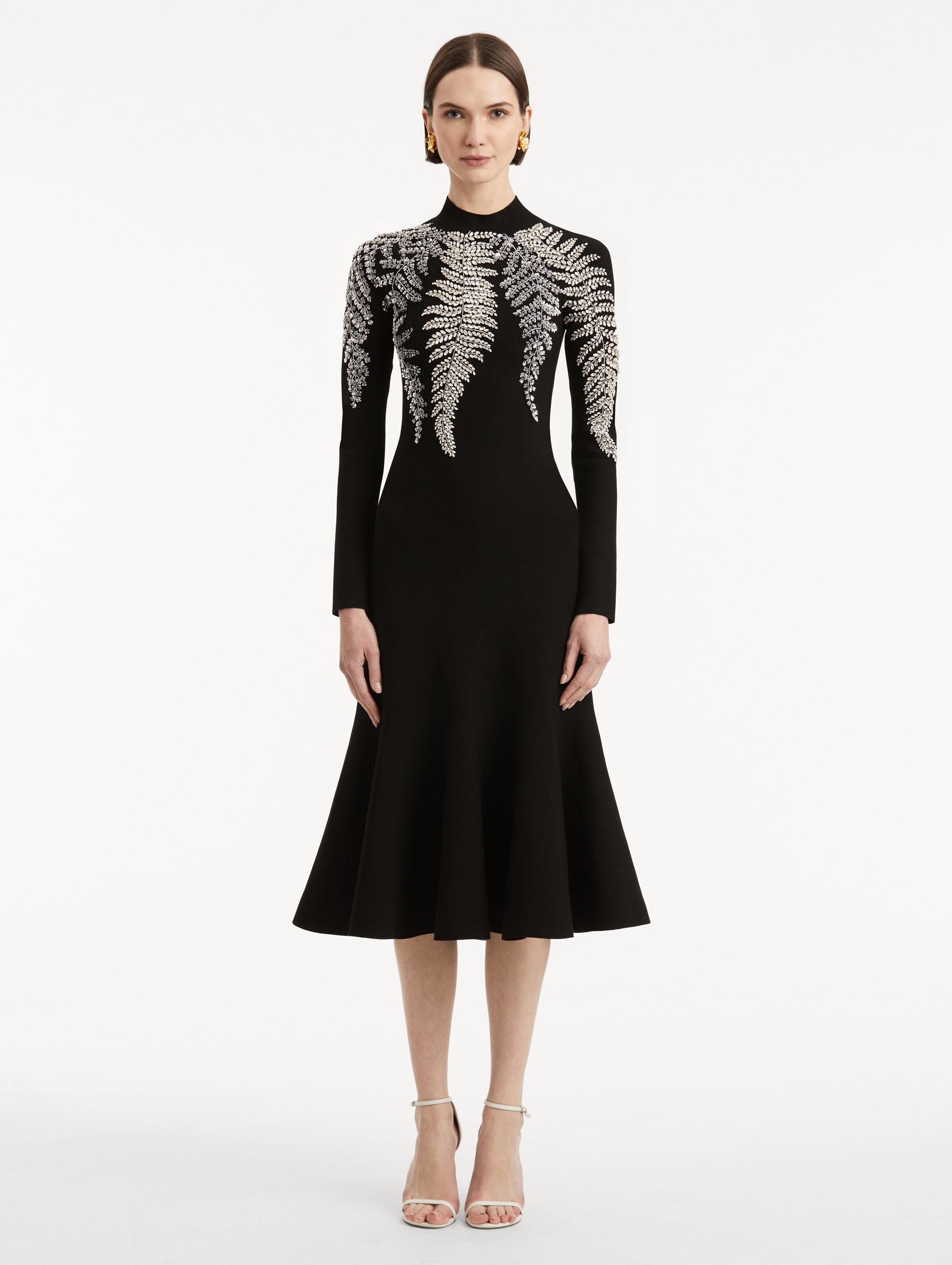 Full body front view image of model wearing a black long sleeve dress with a mock neckline and flared skirt. Features metallic fern embroidery along neck and shoulders. 