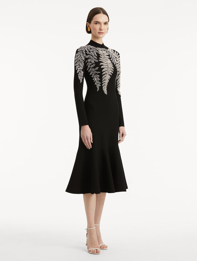 Full body side view image of model wearing a black long sleeve dress with a mock neckline and flared skirt. Features metallic fern embroidery along neck and shoulders. 