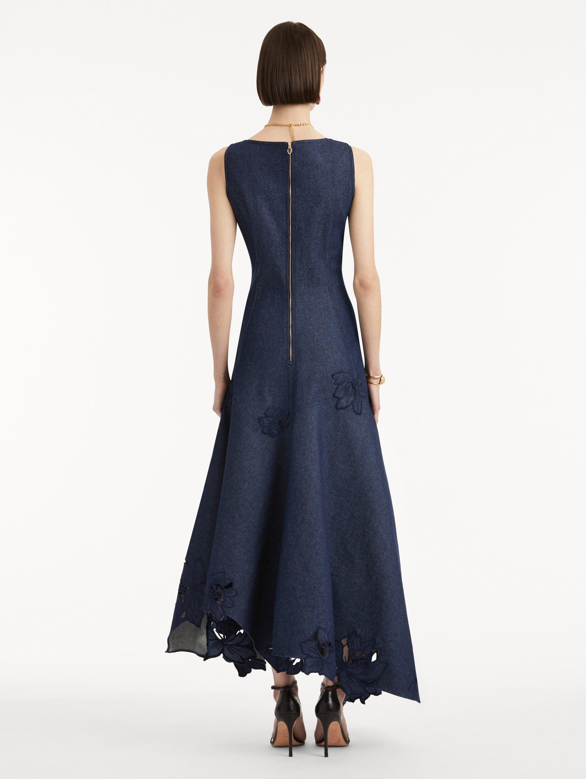 Full body back view image of model wearing a denim dress with peony threadwork cutouts. Features a scoop neckline, gold zipper and asymmetrical hem.