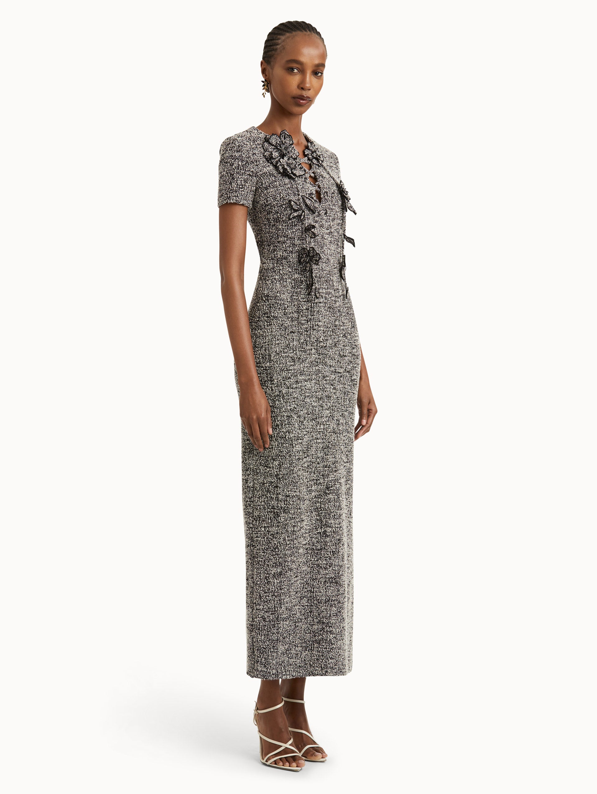 Full body side view of model wearing short sleeve black and ivory tweed dress with floral appliques and lace up neckline. 