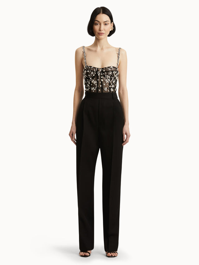 Full body front view image of black jumpsuit with multi-dimensional stones scattered across a tulle bodice and crystal shoulder straps. Features black straight leg pants with side pockets and belt loops. 