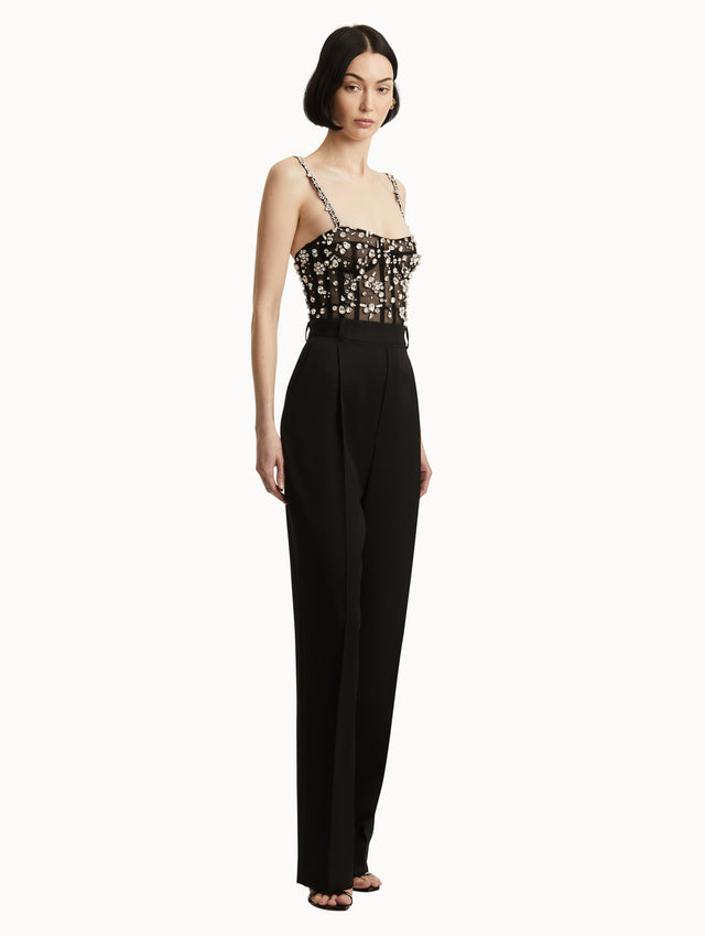 Full body side view image of black jumpsuit with multi-dimensional stones scattered across a tulle bodice and crystal shoulder straps. Features black straight leg pants with side pockets and belt loops. 