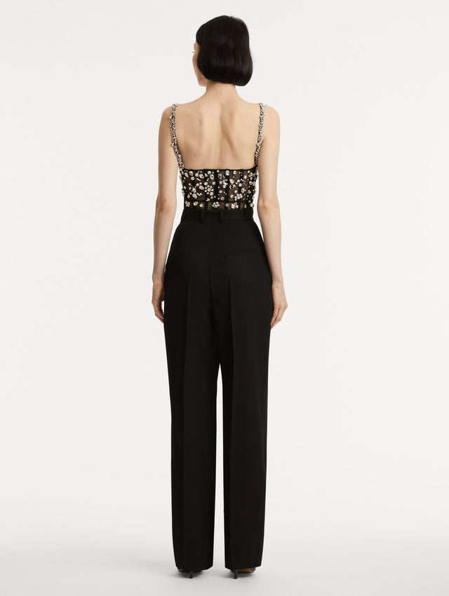 Full body back view image of black jumpsuit with multi-dimensional stones scattered across a tulle bodice and crystal shoulder straps. Features black straight leg pants with side pockets and belt loops. 