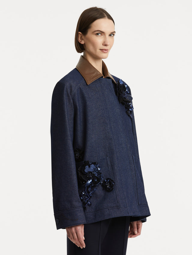 Side view of model wearing dark denim jacket which features navy peony embroidery and a contrasting brown leather spread collar and front patch pockets. 