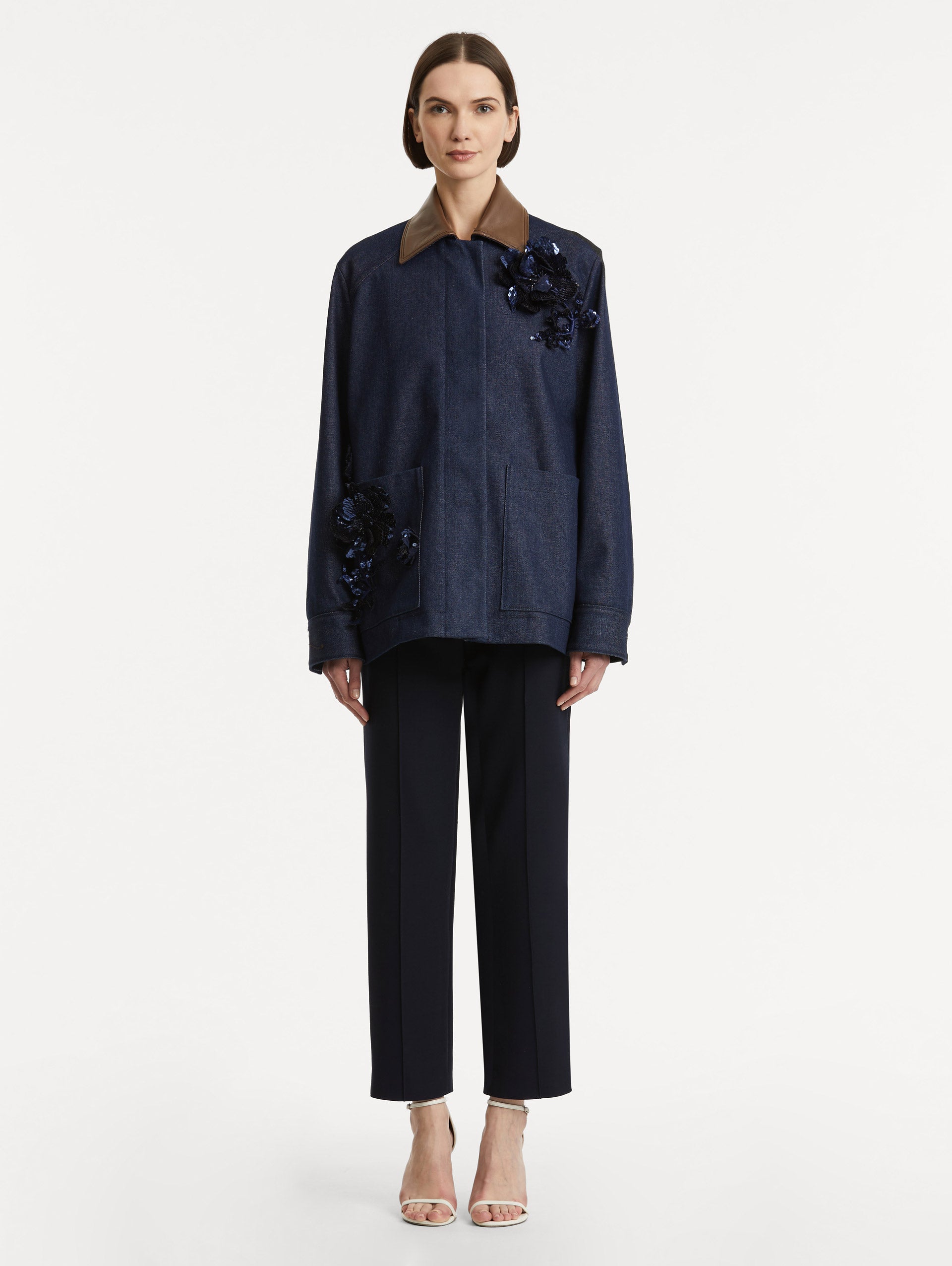 Full body front view of model wearing dark denim jacket which features navy peony embroidery and a contrasting brown leather spread collar and front patch pockets.  