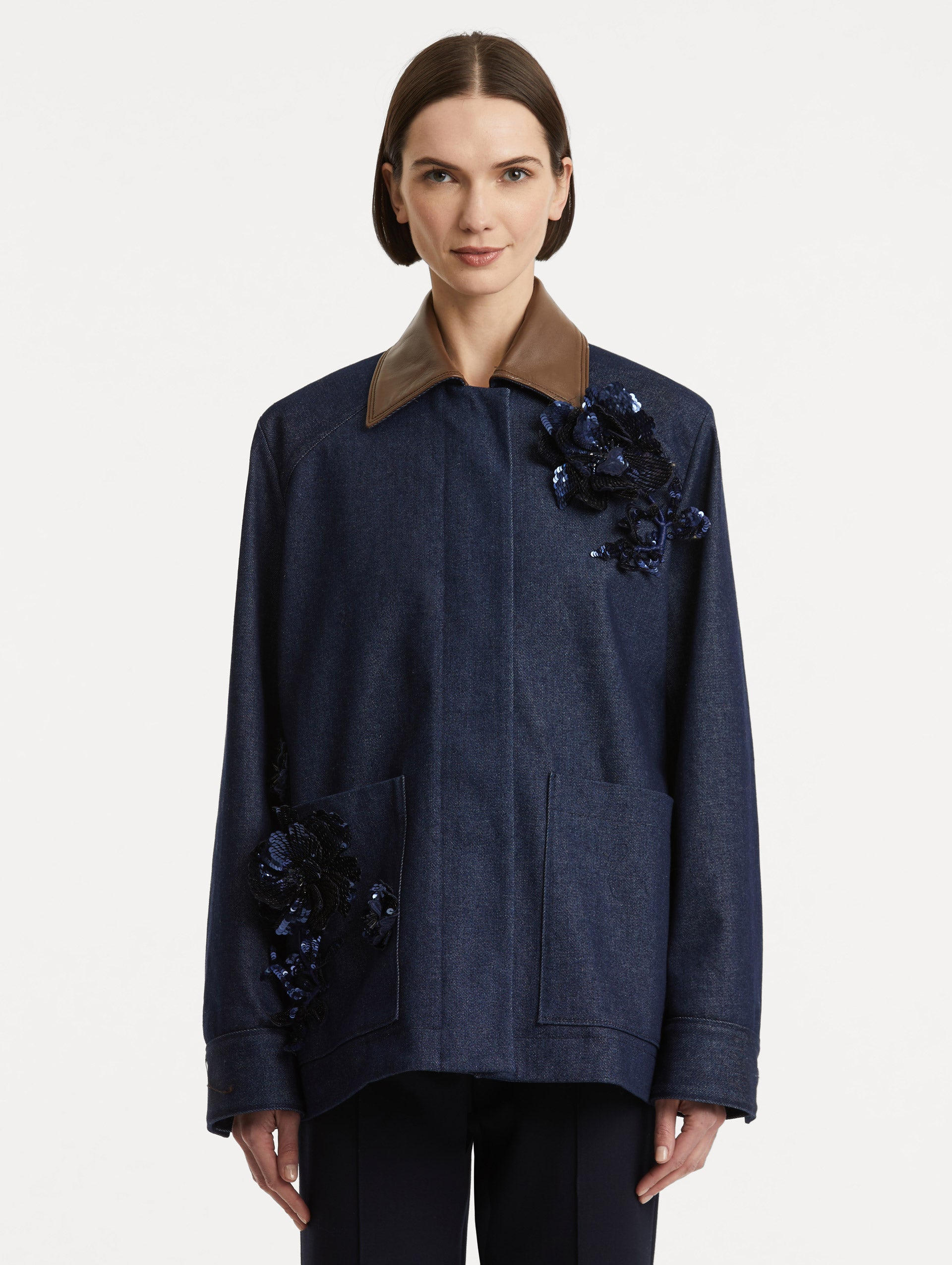 Front view of model wearing dark denim jacket which features navy peony embroidery and a contrasting brown leather spread collar and front patch pockets. 