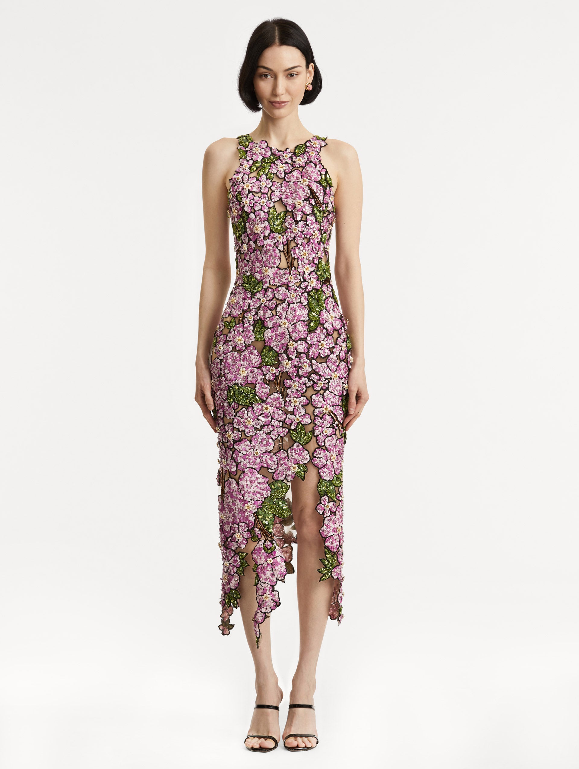 Full body front view of model wearing a sleeveless cocktail dress with embroidered pink flowers and green leaves on a sheer tulle base. While the skirt follows a asymmetrical hem