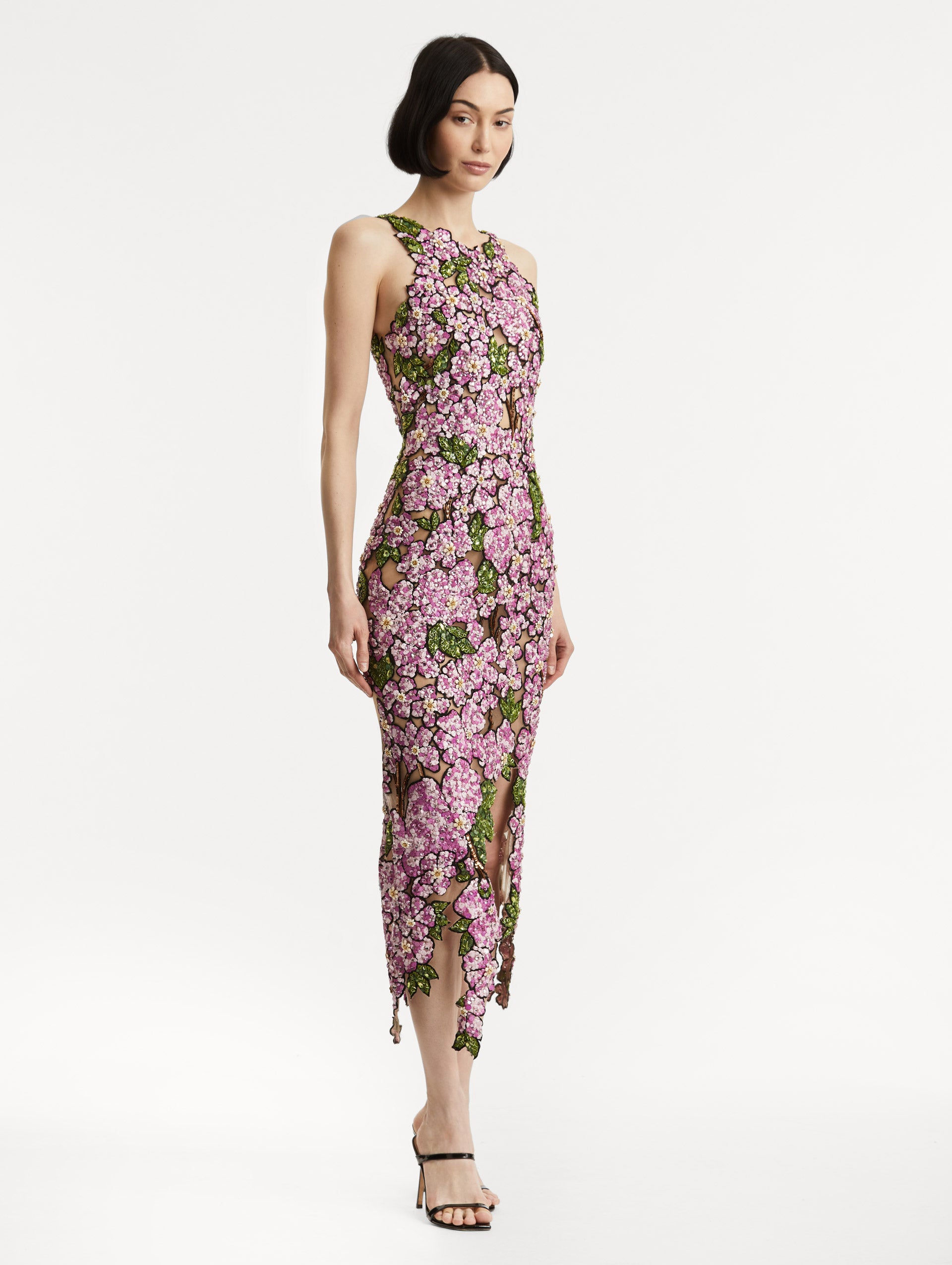 Full body side view of model wearing a sleeveless cocktail dress with embroidered pink flowers and green leaves on a sheer tulle base. While the skirt follows a asymmetrical hem
