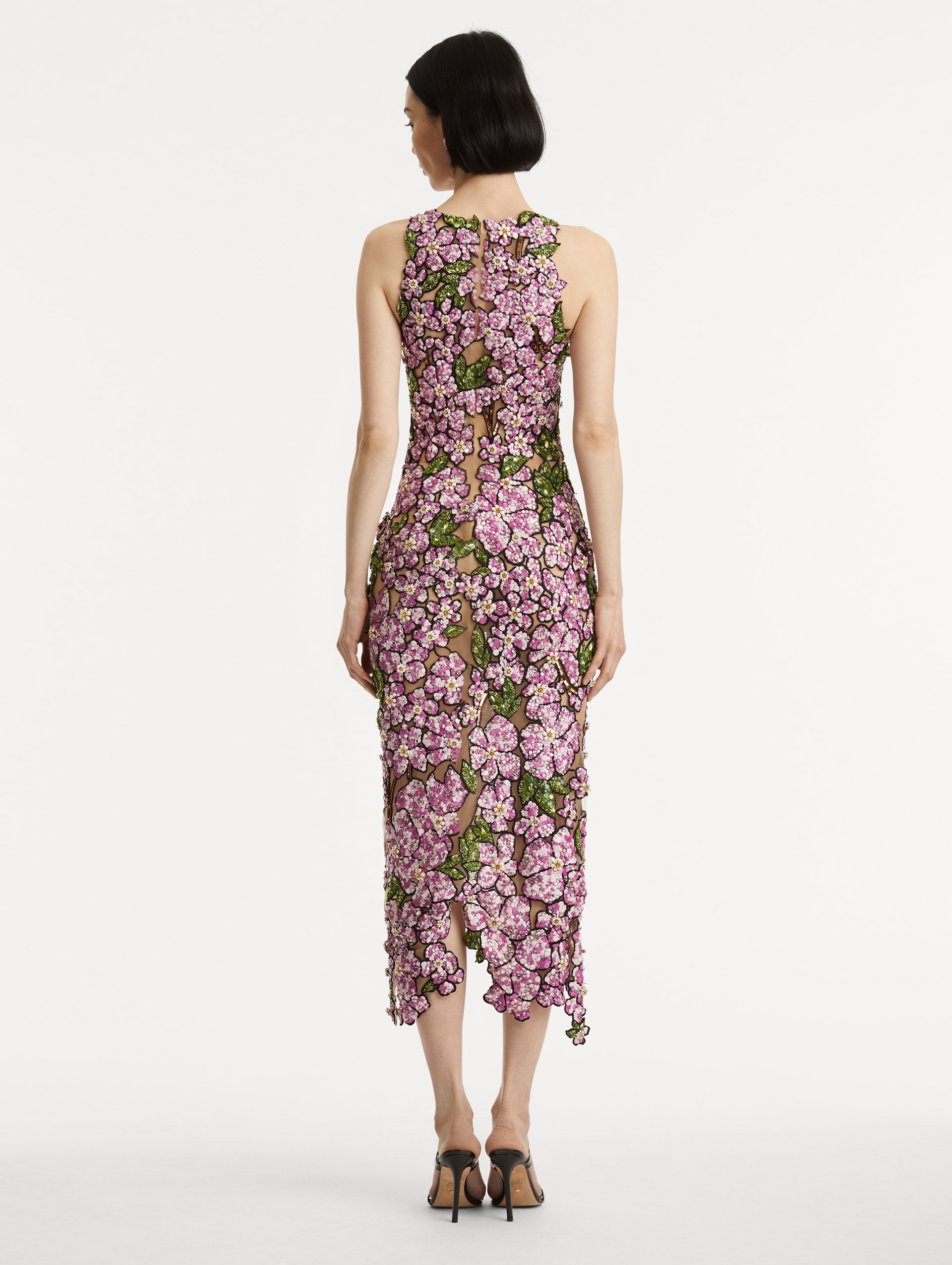 Full body back view of model wearing a sleeveless cocktail dress with embroidered pink flowers and green leaves on a sheer tulle base. While the skirt follows a asymmetrical hem