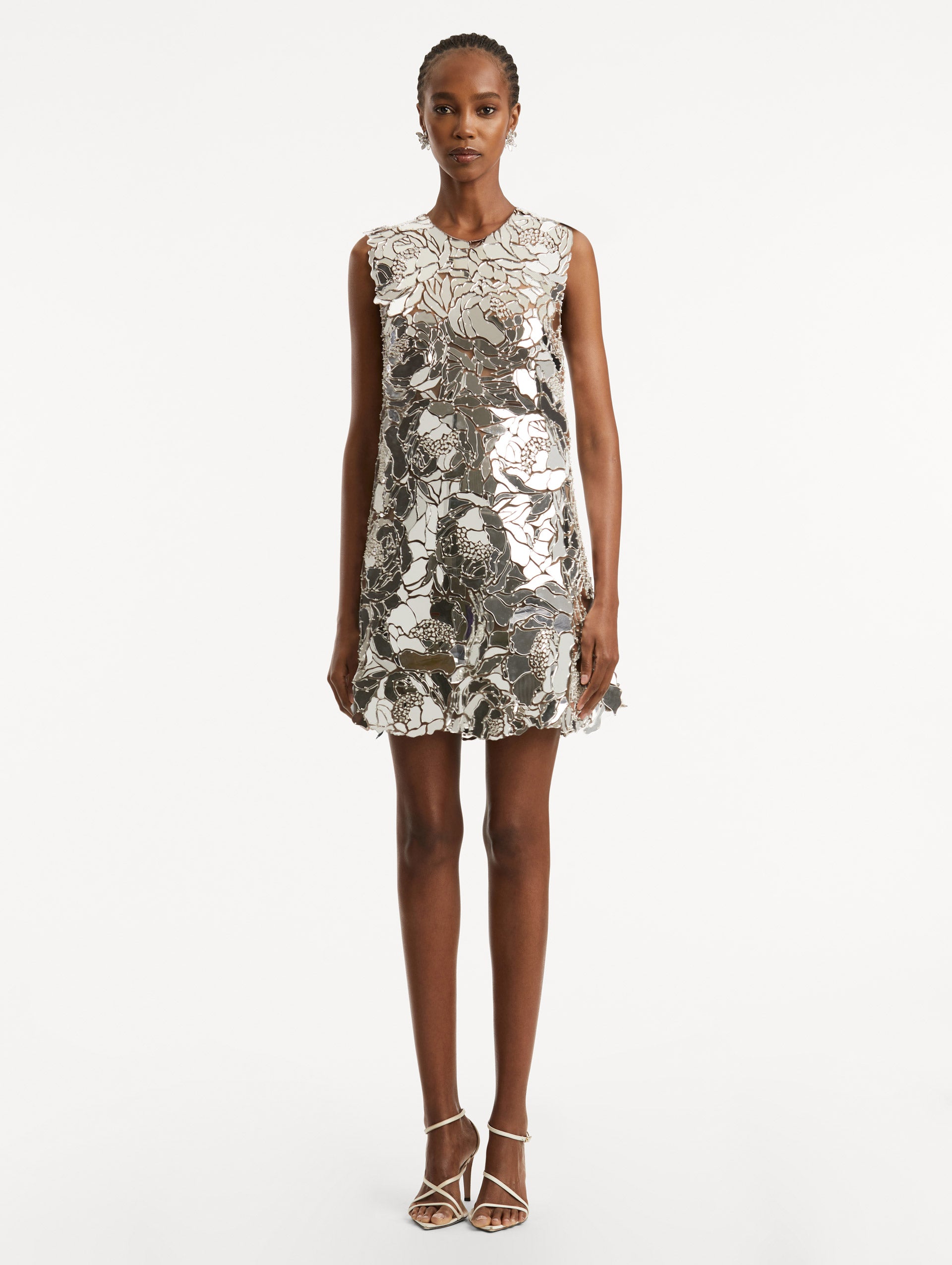 Full body front view of a model wearing a silver shift dress composed of embroidered mirror embellishments in a floral pattern with a jewel neckline.