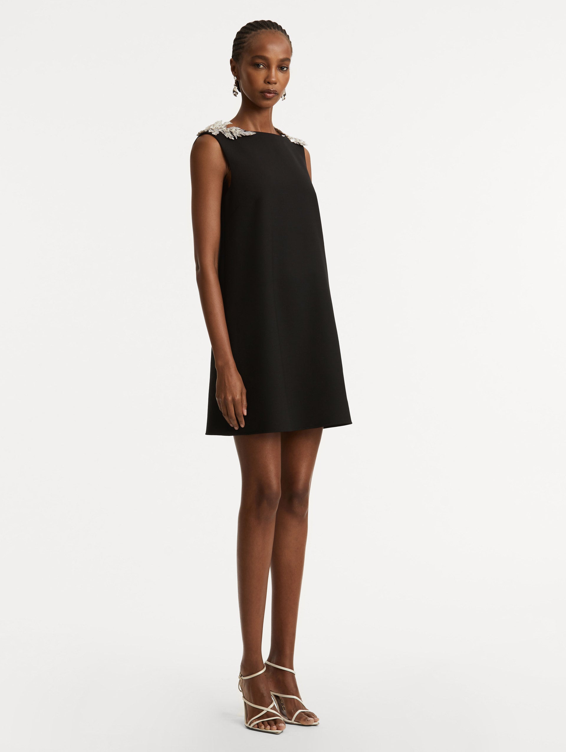 Full body side view of model wearing a black wool minidress in a shift silhouette that features silver sequin fern embroidery on the shoulders. 