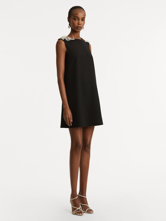 Full body side view of model wearing a black wool minidress in a shift silhouette that features silver sequin fern embroidery on the shoulders. 