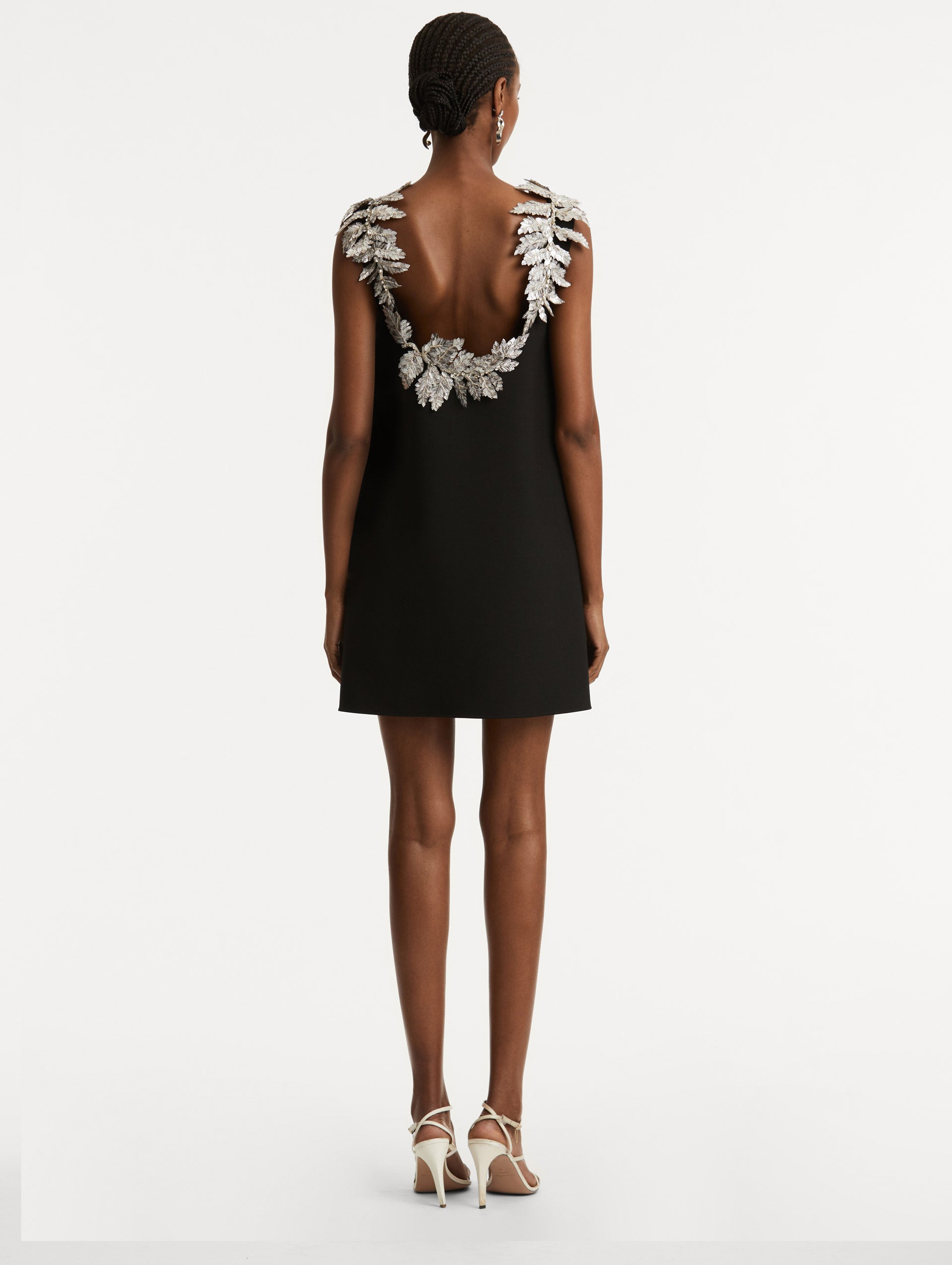 Full body back view of model wearing a black wool minidress in a shift silhouette that features silver sequin fern embroidery following the scoop back. 