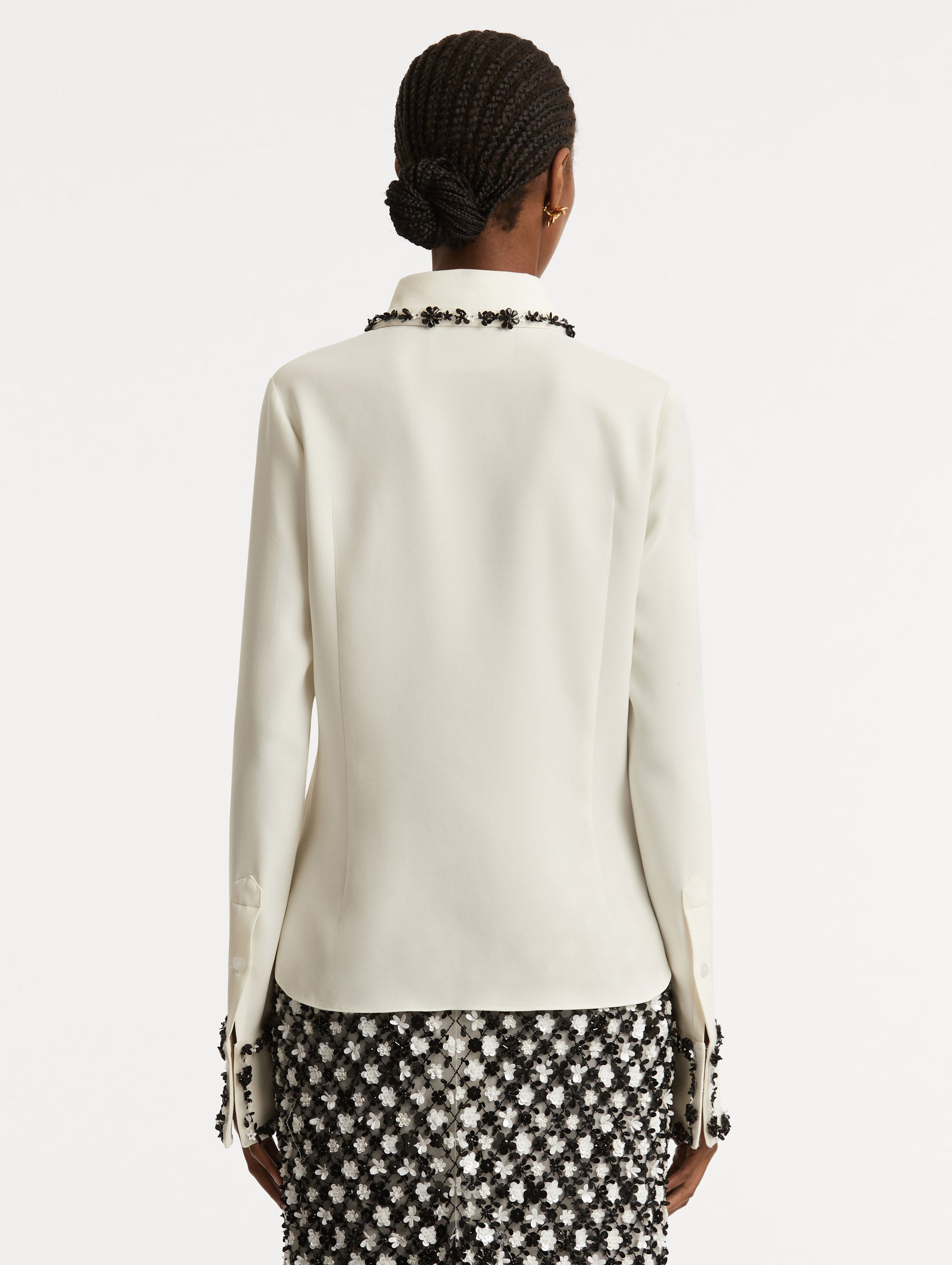 Back view of model wearing a ivory  blouse with black sequin flower embroidery trace the trim with a spread collar.