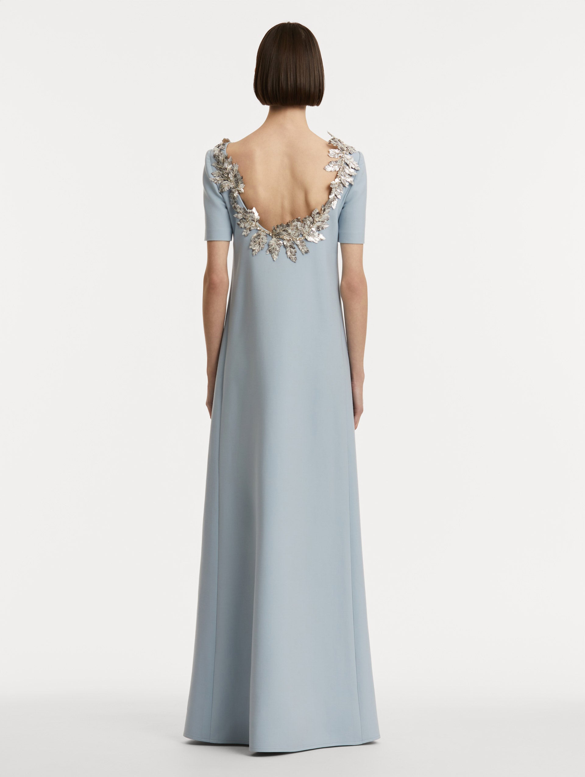 Full body back view of model wearing light blue wool caftan features short sleeves with silver fern embroidery sequins framing the scoop back.