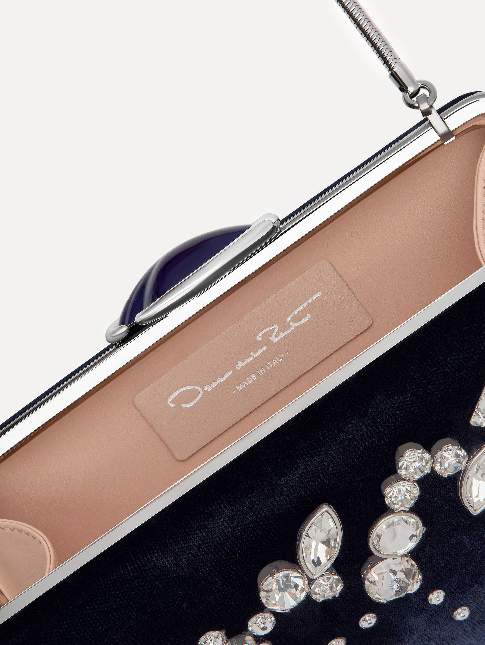 Inside image of Oscar tag inside the clutch featuring the Oscar ‘O’ emblem at the clasp. Clutch with multi-dimensional crystal embroidery and faceted stones scattered across the front canvas. With a detachable chain link shoulder strap