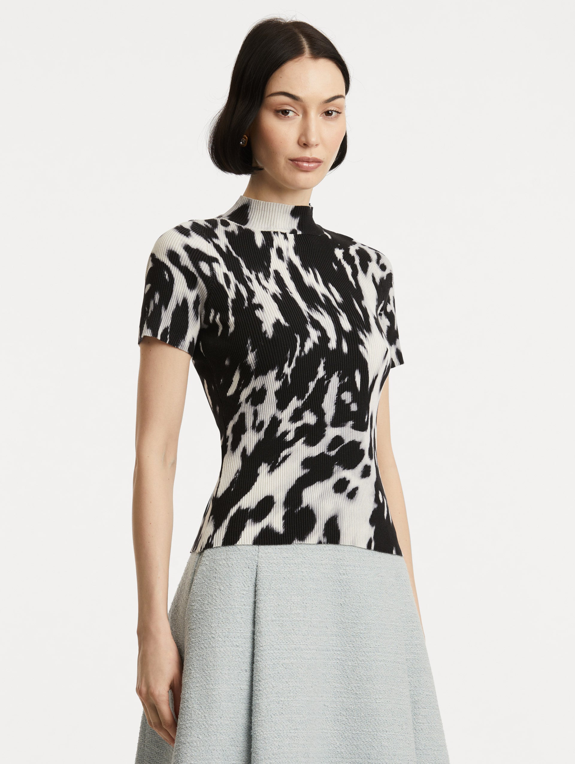 Side view of model wearing short sleeve ribbed knit pullover with a mock neckline and cow print in ecru and black. 