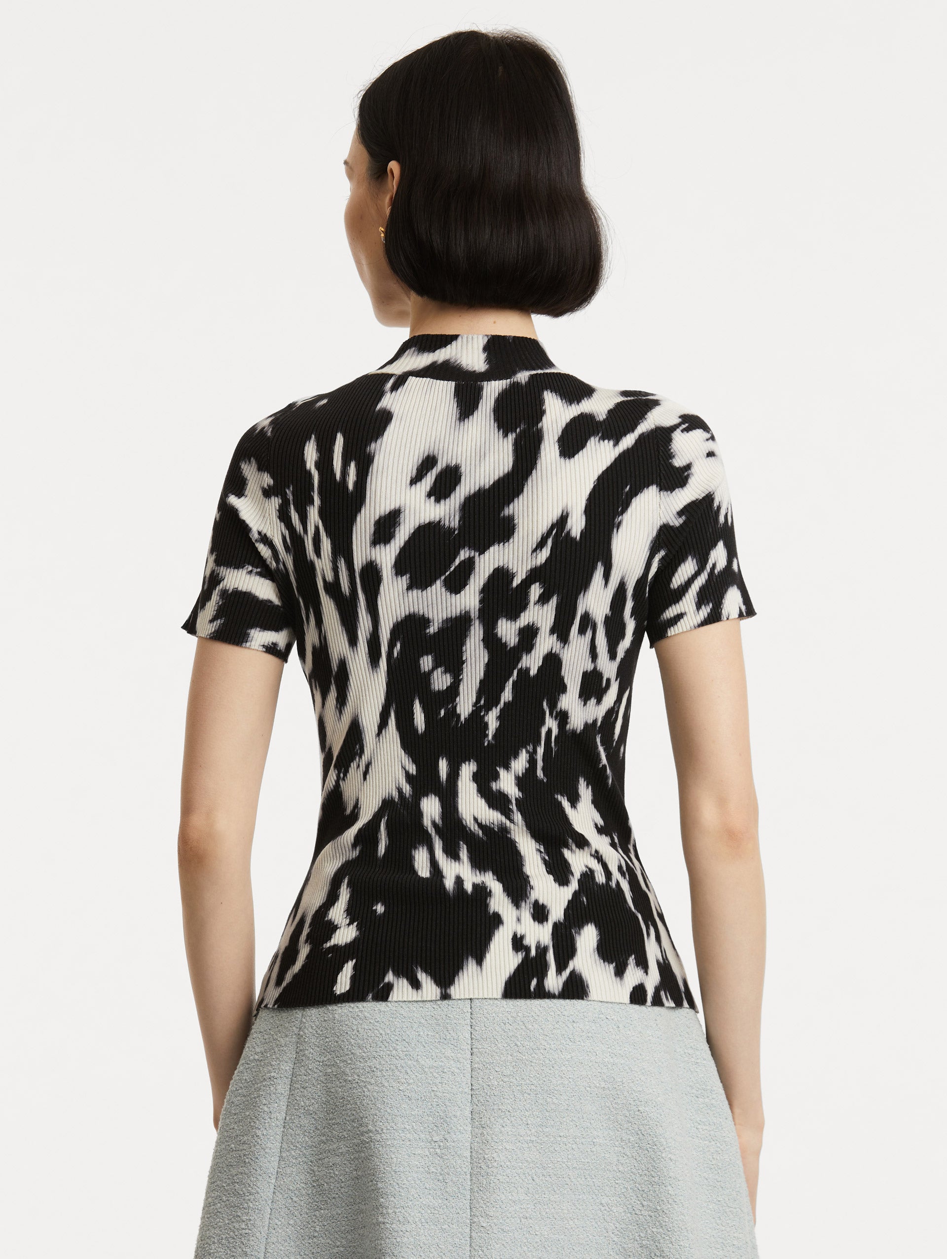 Back view of model wearing short sleeve ribbed knit pullover with a mock neckline and cow print in ecru and black. 