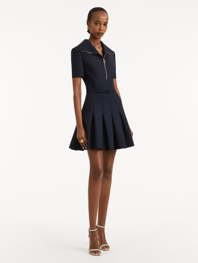 Full body side view of model wearing navy short sleeve mini dress with gold zipper, spread collar, matching self-belt, and pleated skirt. 