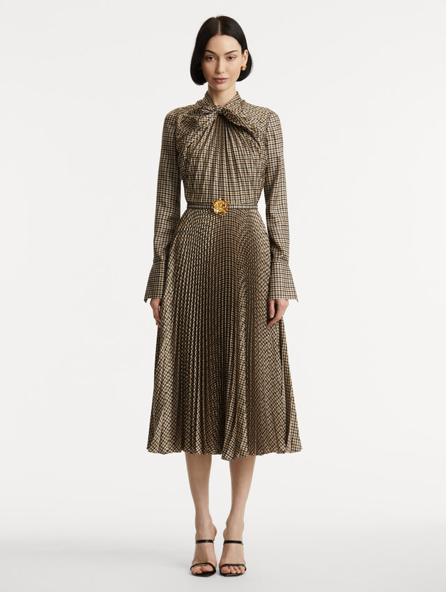 Full body front view of model wearing a multiple brown colored dress with a long sleeves and a neck tie. Dress also features pleating and a matching self-belt with a gold peony at the center. 