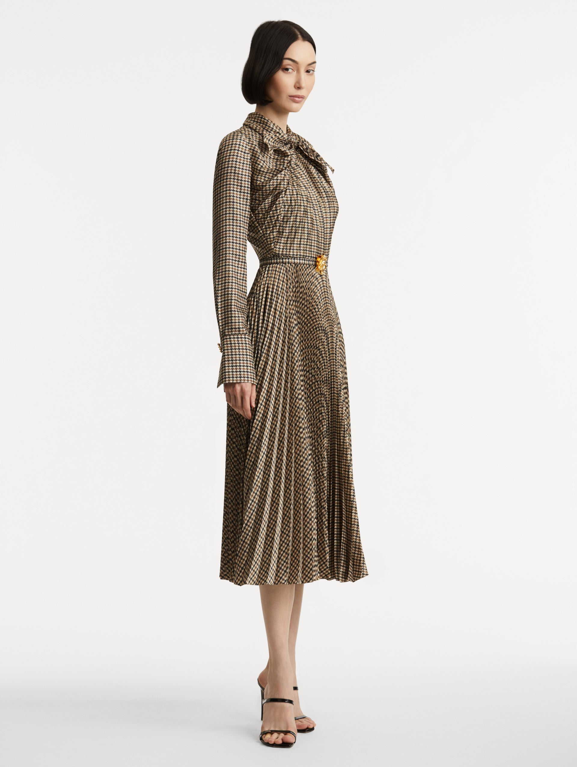Full body side view of model wearing a multiple brown colored dress with long sleeves and a neck tie. Dress also features pleating and a matching self-belt with a gold peony at the center. 