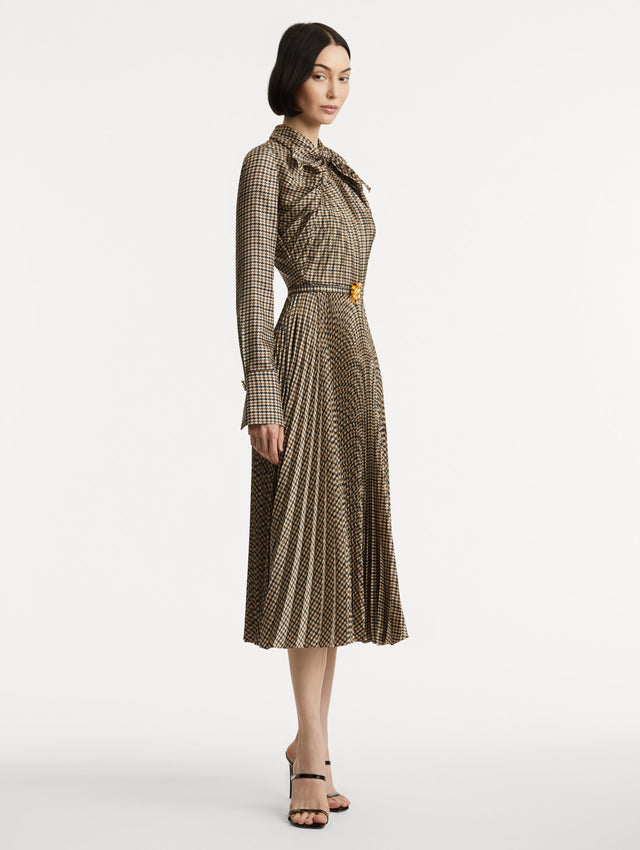 Full body side view of model wearing a multiple brown colored dress with long sleeves and a neck tie. Dress also features pleating and a matching self-belt with a gold peony at the center. 