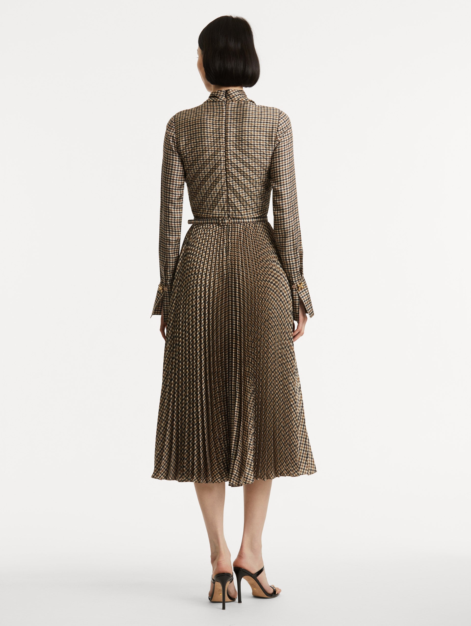 Full body back view of model wearing a multiple brown colored dress a with long sleeves. Dress also features pleating and a matching self-belt with a gold peony at the center. 