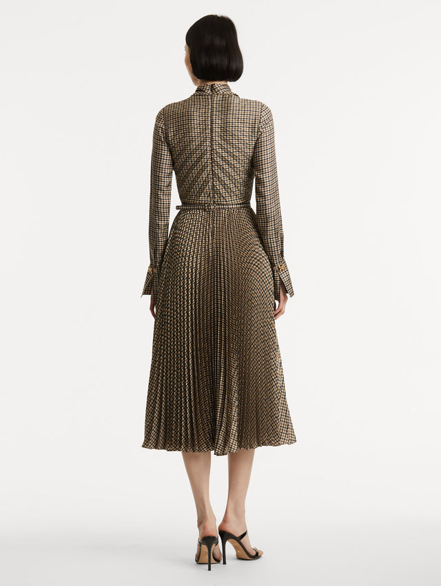 Full body back view of model wearing a multiple brown colored dress a with long sleeves. Dress also features pleating and a matching self-belt with a gold peony at the center. 