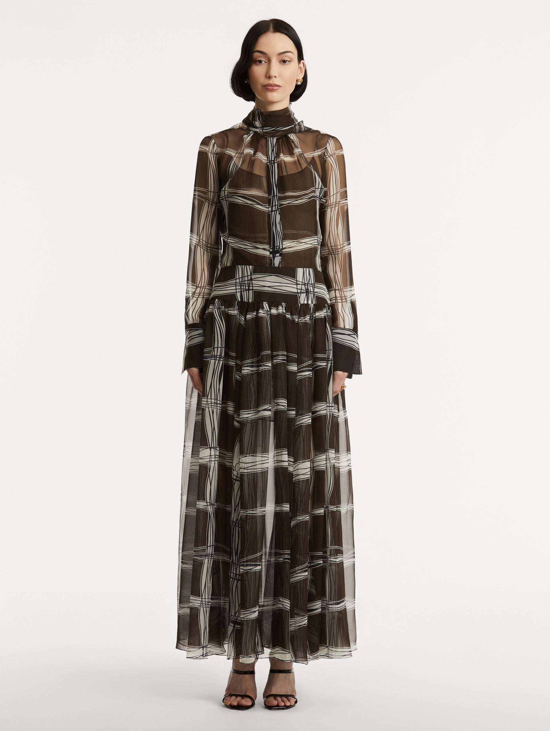 Full body front view of silk chiffon dress which features a brown base with a hand-drawn plaid pattern. Dress has long sleeves and a mock neckline with a self-tie closure. 