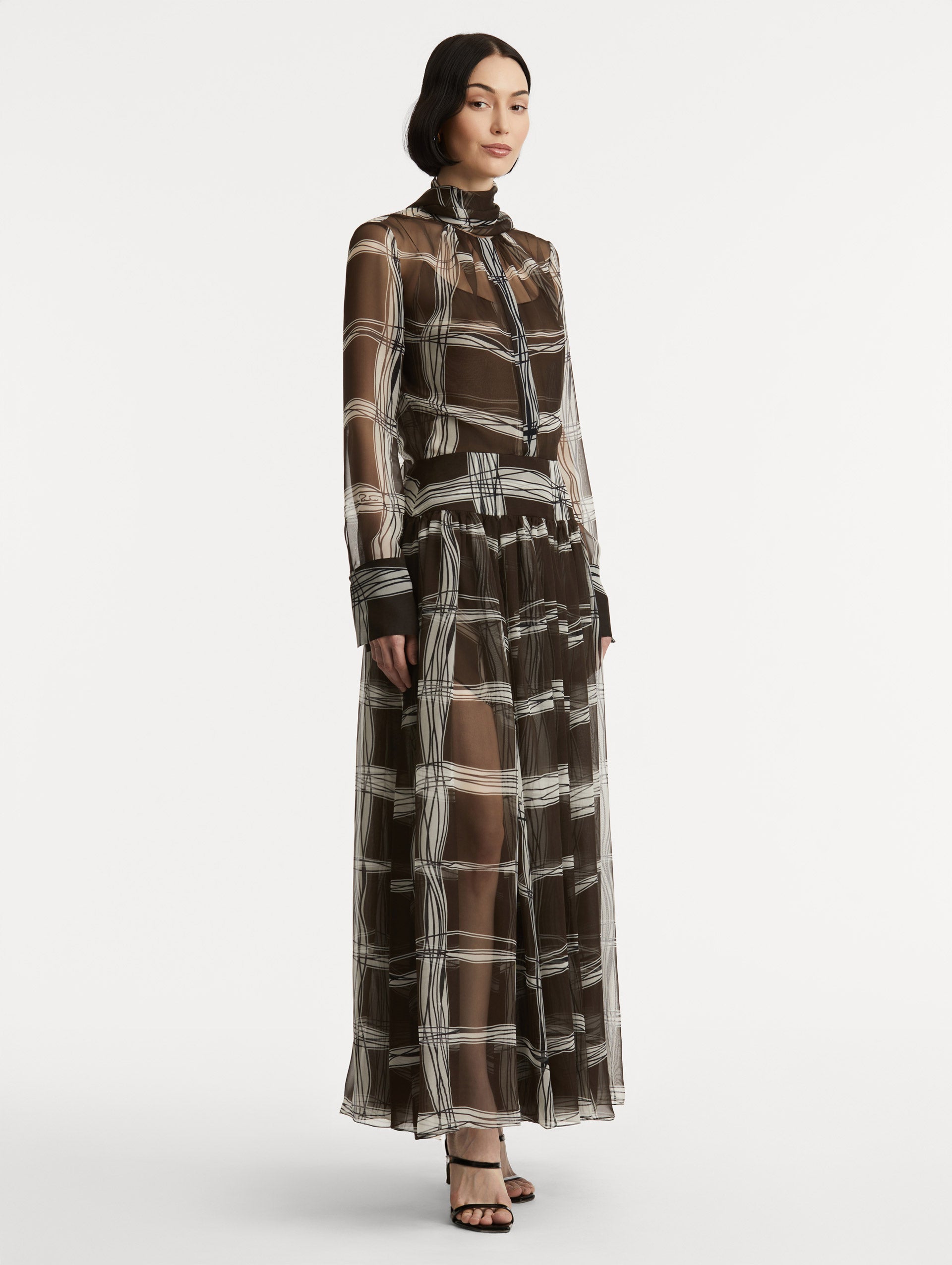 Full body side view of silk chiffon dress which features a brown base with a hand-drawn plaid pattern. Dress has long sleeves and a mock neckline with a self-tie closure. 