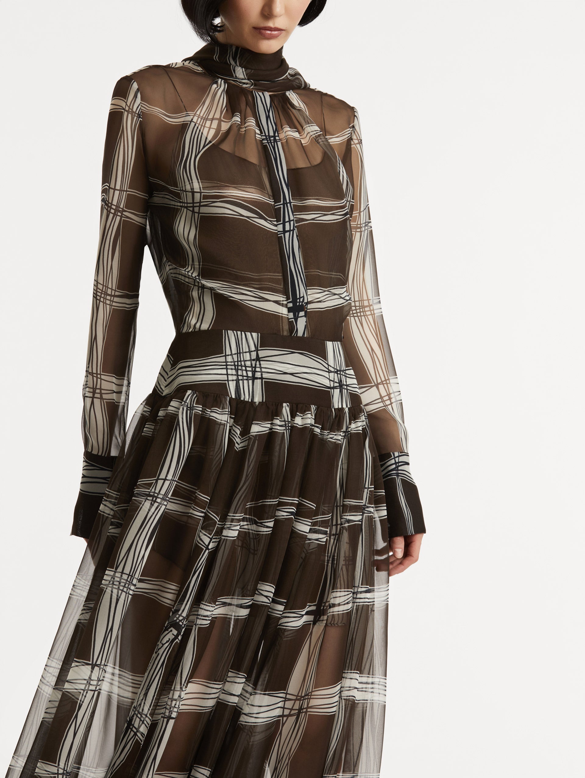 Zoomed in front view of silk chiffon dress which features a brown base with a hand-drawn plaid pattern. Dress has long sleeves and a mock neckline with a self-tie closure and high waistband.